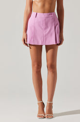 A woman is wearing a high-waisted, pastel pink mini skirt with a subtle checkered pattern. The skirt features belt loops and vertical seam details running down the front. She is also wearing white high-heeled sandals with ankle straps. The image focuses on her lower torso and legs against a plain, light background.
